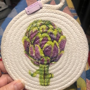 May include: A white, round, crocheted pot holder with a green and purple painted artichoke design. The pot holder has a loop handle and a "HANDMADE" tag.