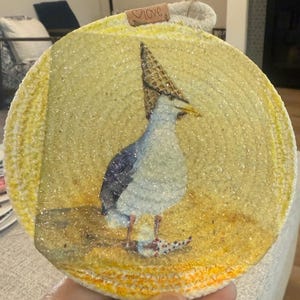 May include: Round, yellow ornament with a seagull wearing a waffle cone hat. The seagull is painted in shades of blue, gray, and white. The ornament has a rope hanger and a small tag that says "love".