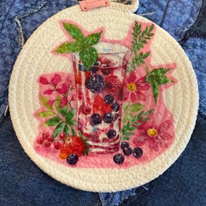 May include: Round white coaster with a watercolor painting of a refreshing drink with berries and mint. The coaster has a pink border and a loop for hanging. The drink includes strawberries, blueberries, and raspberries.