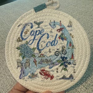May include: Round white trivet with a rope edge, featuring a map of Cape Cod in blue, with illustrations of a lobster, lighthouse, and bicycle. The words "Cape Cod" are written in blue script.