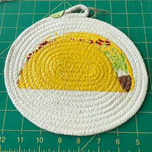 May include: A round, white, woven trivet with a yellow taco design. The taco is painted with yellow, red, green, and brown details. A loop of rope is attached for hanging. The trivet is approximately 8 inches in diameter.