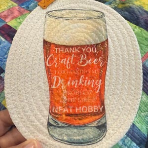 May include: A round, white woven coaster with a beer glass design. The glass is filled with amber-colored beer and has the text "Thank You, Craft Beer for Making My Drinking Problem Look Like a Neat Hobby."