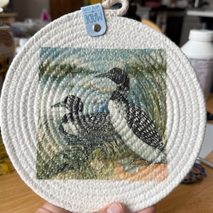 May include: Round, off-white woven trivet featuring a watercolor image of two loons in a natural setting. The loons are black and white with detailed feather patterns. A small blue tag with the words "MADE WITH LOVE" is attached.