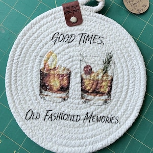 May include: A round, white coiled rope coaster featuring a printed graphic of two cocktails with the text "GOOD TIMES, OLD FASHIONED MEMORIES." A brown leather tab labeled "hand made with love" is attached at the top with a silver metal rivet.