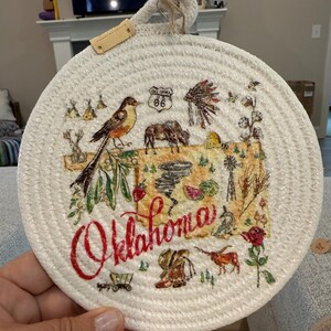 May include: Round, woven trivet with a painted Oklahoma design. The design includes a bird, Route 66 sign, bison, and the word "Oklahoma" in red script. The trivet has a loop for hanging and a small leather tag.