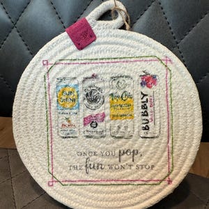May include: Round white woven trivet with a printed design of four beverage cans. The cans are labeled "Sun Shine", "Waves", "Top Chic", and "Bubbly". Text reads "Once you pop, the fun won't stop". A pink tag is attached.