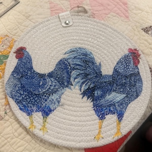 May include: Round white woven trivet featuring two blue roosters with red combs and yellow feet. The trivet has a loop for hanging and the text "MADE WITH LOVE" on a small tag.