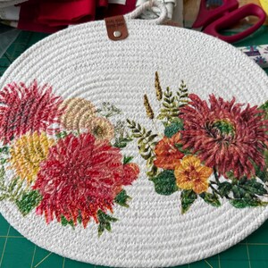 May include: A round, white woven trivet with a floral design. The trivet features a cluster of red, yellow, and orange flowers with green leaves. A small leather tag is attached to the top with a loop for hanging.