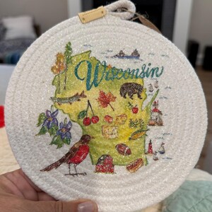 May include: Round, cream-colored trivet with a detailed map of Wisconsin. The map is green and features illustrations of local landmarks, a red robin, and the word "Wisconsin" in teal script. A small leather tag is attached.