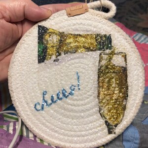 May include: Round, white coaster with a champagne bottle pouring into a glass, both covered in gold glitter. The word "cheers!" is written in blue cursive. A small brown tag hangs from the top.