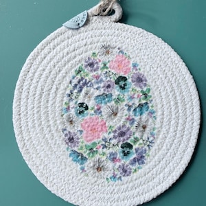 May include: Round white woven trivet with a floral design in the center. The floral design is in the shape of an egg and features pink, purple, blue, and green flowers. A small tag is attached to the top with a natural fiber cord.