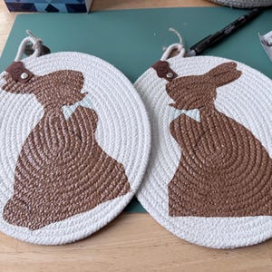 May include: Two white, round trivets with a brown bunny design. Each trivet is made of woven white rope with a brown bunny silhouette in the center. They have a small leather loop for hanging.