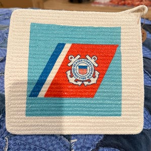 May include: Square, off-white coaster with a light blue and red design featuring the United States Coast Guard emblem. The coaster has a light blue background with a diagonal red stripe and a blue stripe. The emblem includes the words "United States Coast Guard 1790".