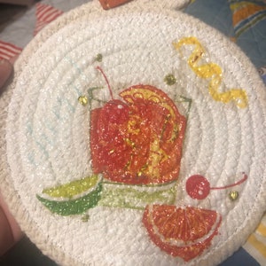 May include: A round, white woven coaster with a painted cocktail design. The cocktail features a red cherry, orange slice, and lime wedge. The coaster has a loop for hanging and a small tag that says "HANDMADE".
