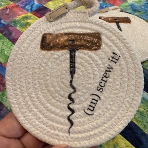 May include: White, round coaster with a rope design and a corkscrew illustration. The coaster has the phrase "(un) screw it!" printed on it. A small tag is attached to the top with a rope loop.