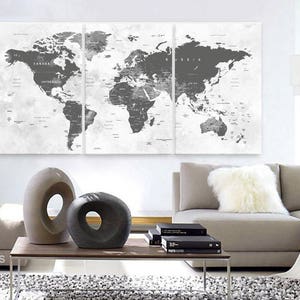 May include: A black and white world map print on canvas, divided into three panels. The map is displayed on a white wall above a gray couch with a white throw pillow. The couch is in a living room setting with a coffee table and two decorative sculptures.