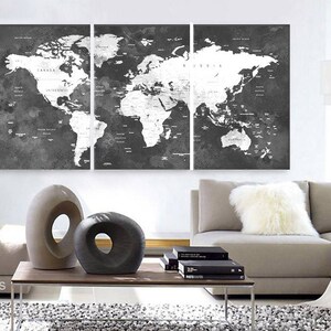 May include: A black and white world map print with a distressed, vintage look. The map is divided into three panels and features a white outline of the continents and countries. The map is displayed on a wall above a gray couch with a white throw pillow.
