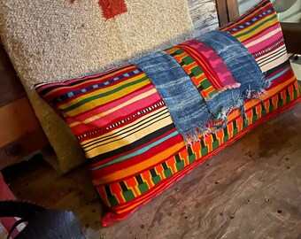 May include: A colorful striped pillow with a denim patch and frayed edges. The pillow is resting on a wooden surface with a red cross on a beige fabric in the background.