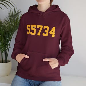 Op de afbeelding: Bordeauxkleurig hoodie met een groot geel nummer "55734" gedrukt op de voorkant.
