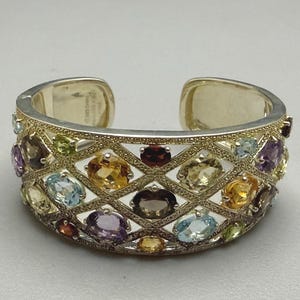 Puede incluir: Una pulsera de brazalete ancha con una estructura de celosía plateada con un acabado dorado texturizado. El diseño calado está engastado con numerosas piedras preciosas ovaladas de varios colores, incluyendo citrino, granate, amatista y topacio azul.