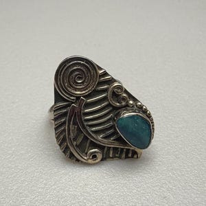 May include: A silver ring featuring an asymmetrical, textured metal face with a prominent spiral design and a single, teardrop-shaped teal stone set on the right side. The metal surface displays deep grooves and raised lines throughout the piece.