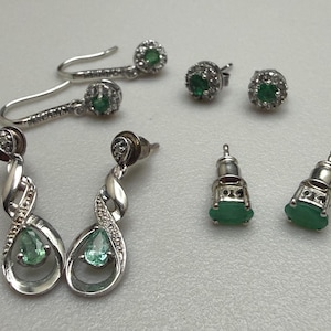May include: A collection of three pairs of silver-toned earrings featuring green stones. One pair consists of drop earrings with pear-shaped green stones, another has hook-style earrings with round green stones, and the third pair are stud earrings.
