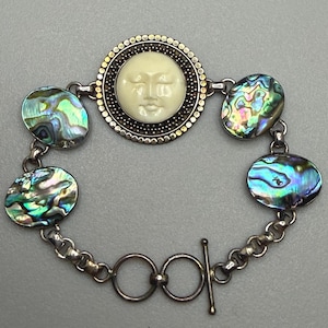 May include: A bracelet featuring a central circular charm with a cream-colored carved moon face surrounded by a beaded metal border, alternating with four oval iridescent abalone shell cabochons, all connected by silver-toned metal links and a toggle clasp.