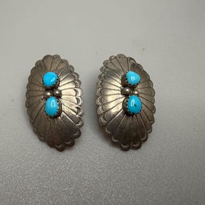 Könnte beinhalten: Paar silberfarbene ovale Ohrringe. Jeder Ohrring hat zwei leuchtend türkisfarbene Steine, die in einem dekorativen, gewellten Design gefasst sind. Die Ohrringe haben einen Vintage- oder Südwest-Stil.