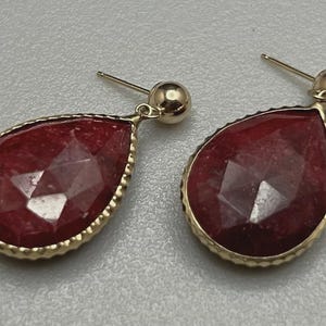 May include: A pair of gold-colored drop earrings with faceted, teardrop-shaped, deep red gemstones. The gemstones are set in a gold-colored bezel with a textured edge. The earrings have a post and ball closure.