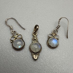 K&ouml;nnte beinhalten: Ein silbernes Schmuckset mit einem Anh&auml;nger und Ohrringen. Jedes St&uuml;ck hat einen runden, irisierenden Stein in einem dekorativen Silberrahmen. Die Ohrringe haben einen Hakenverschluss.