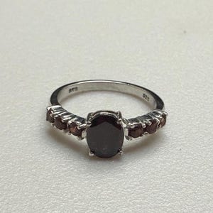 Op de afbeelding: Een zilveren ring met een grote, ovaal geslepen donkerrode edelsteen in het midden, geflankeerd door drie kleinere ronde donkerrode edelstenen aan weerszijden van de ringband. De binnenkant van de zilveren ring is gestempeld met 925.