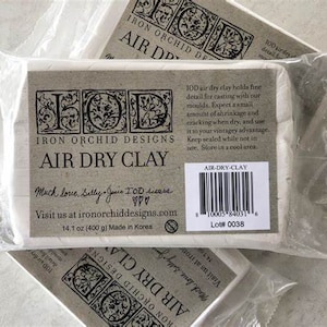 May include: A package of Iron Orchid Designs Air Dry Clay, a white clay that is 14.1 ounces (400 grams) and made in Korea. The package has a black and white label with the company logo and product information. The label also includes a barcode and lot number.