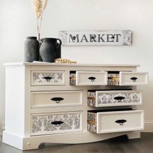 May include: White dresser with six drawers, some open, revealing patterned interiors. Black handles and a "MARKET" sign above. Two dark grey vases and a decorative vase with dried plants sit on top. The dresser is approximately 102 cm wide.