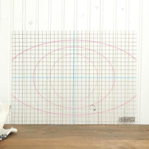 May include: A clear, rectangular cutting mat with a grid pattern and pink spiral design. Blue lines intersect at the center. The mat has a logo in the bottom right corner and is placed on a wooden surface.
