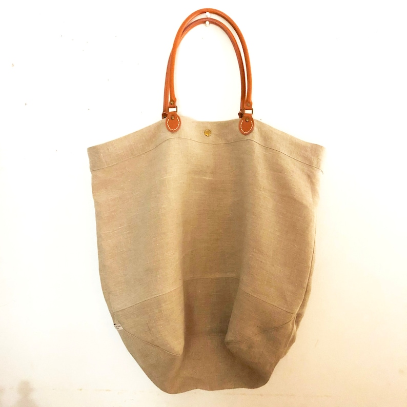 Puede incluir: Un bolso tote grande de lino beige con asas de cuero marr&oacute;n. El bolso tiene un dise&ntilde;o simple y cl&aacute;sico y es perfecto para llevar los art&iacute;culos esenciales del d&iacute;a a d&iacute;a.