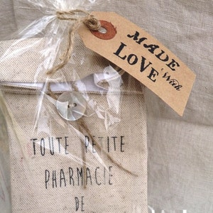 Op de afbeelding: Een kleine, beige canvas tas met de tekst "TOUTE PETITE PHARMACIE DE VOYAGE" gedrukt op de voorkant. De tas is verpakt in doorzichtig plastic en vastgebonden met touw. Een bruin label met de tekst "MADE with LOVE" is aan het touw bevestigd.