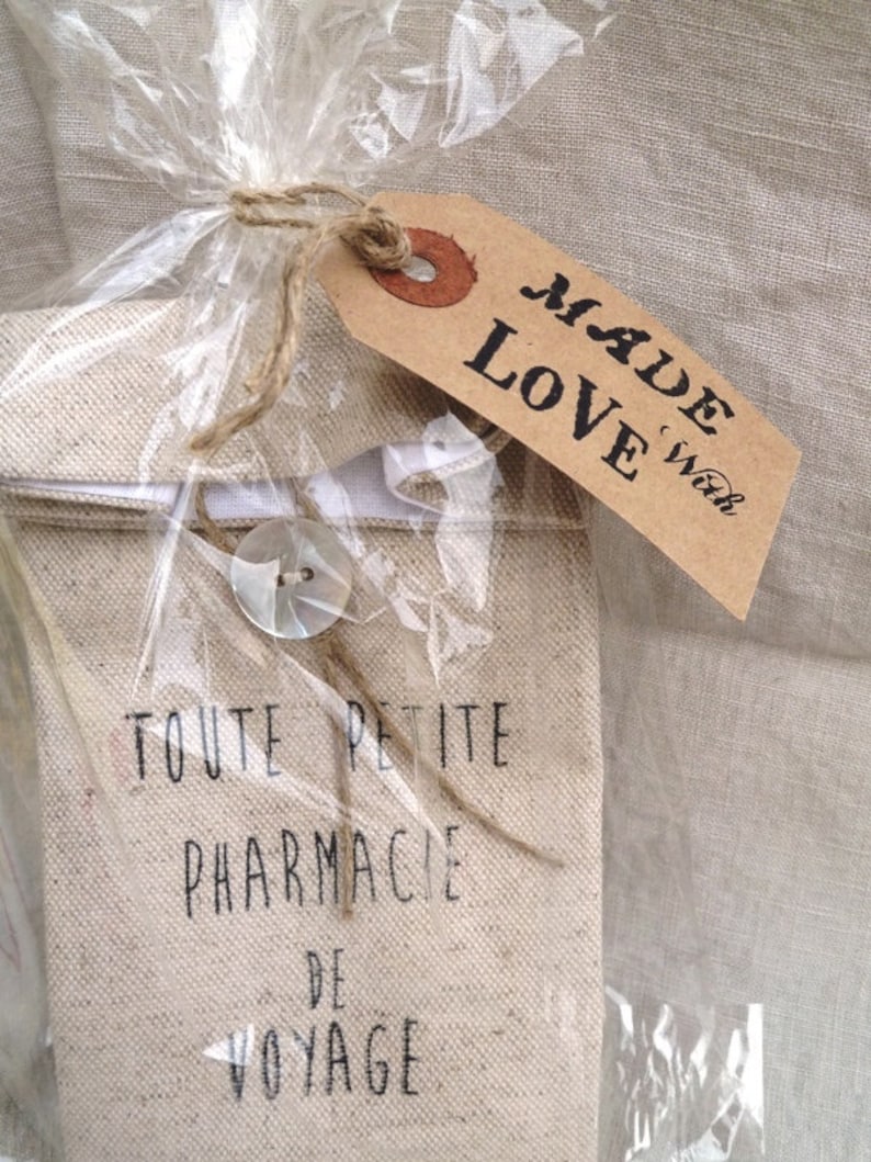 Op de afbeelding: Een kleine, beige canvas tas met de tekst "TOUTE PETITE PHARMACIE DE VOYAGE" gedrukt op de voorkant. De tas is verpakt in doorzichtig plastic en vastgebonden met touw. Een bruin label met de tekst "MADE with LOVE" is aan het touw bevestigd.