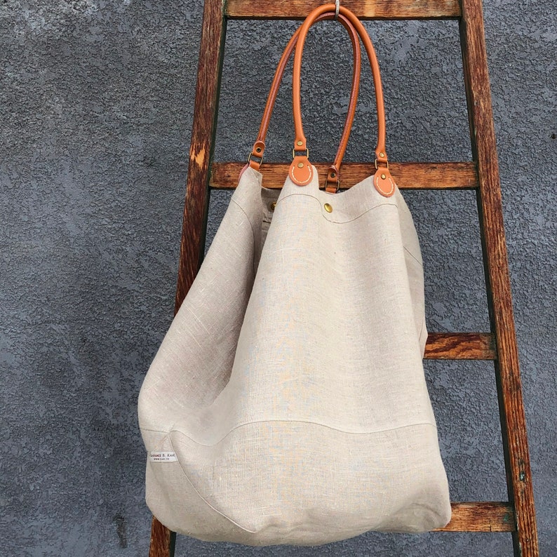 Puede incluir: Un bolso tote grande de lino de color blanco roto con asas de cuero marr&oacute;n y herrajes de lat&oacute;n. El bolso est&aacute; colgado de una escalera de madera.