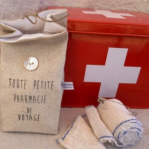 Op de afbeelding: Een klein, beige linnen tasje met de tekst "TOUTE PETITE PHARMACIE DE VOYAGE" erop gedrukt. Het tasje ligt naast een rode metalen EHBO-doos met een wit kruis op de voorkant. Er liggen ook witte gaasverbanden met blauwe rand.