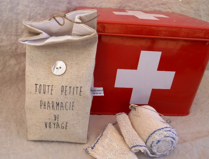 Op de afbeelding: Een klein, beige linnen tasje met de tekst "TOUTE PETITE PHARMACIE DE VOYAGE" erop gedrukt. Het tasje ligt naast een rode metalen EHBO-doos met een wit kruis op de voorkant. Er liggen ook witte gaasverbanden met blauwe rand.