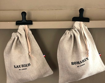 Puede incluir: Dos bolsas de lino beige con cordón y texto negro. La bolsa de la izquierda dice "Laurier du Jardin" y la bolsa de la derecha dice "Romarin du Jardin".