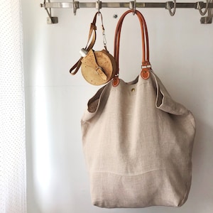 Puede incluir: Un bolso tote grande de lino beige con asas y correas de cuero marr&oacute;n. El bolso est&aacute; colgado en un estante de metal con un peque&ntilde;o bolso redondo de cuero marr&oacute;n con una hebilla de metal colgando al lado.