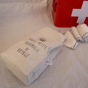 Op de afbeelding: Een kleine witte canvas tas met de tekst "Toute Petite Pharmacie De Voyage" erop gedrukt. De tas ligt op een wit stoffen oppervlak naast een rode EHBO-doos met een wit kruis.