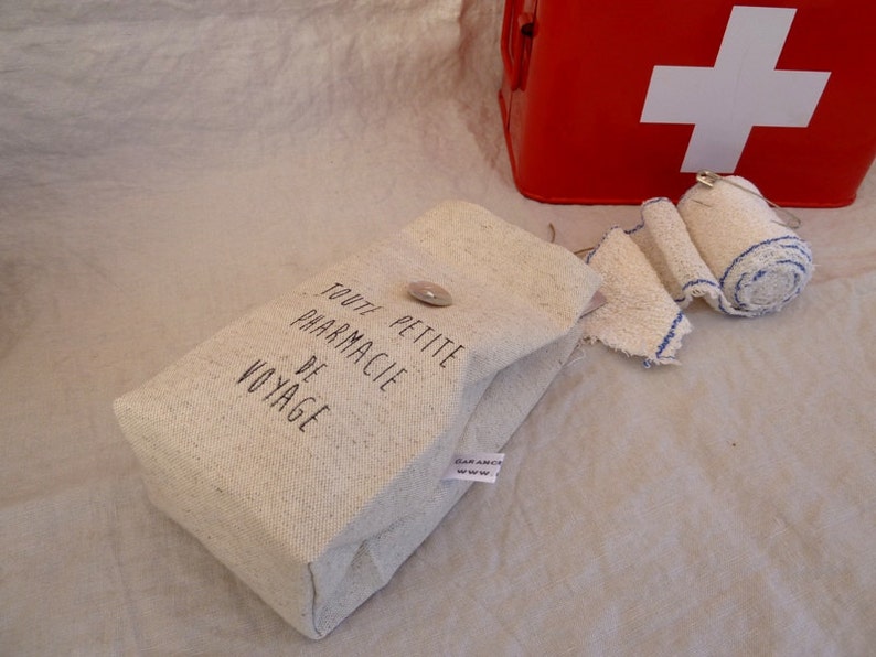 Op de afbeelding: Een kleine witte canvas tas met de tekst "Toute Petite Pharmacie De Voyage" erop gedrukt. De tas ligt op een wit stoffen oppervlak naast een rode EHBO-doos met een wit kruis.