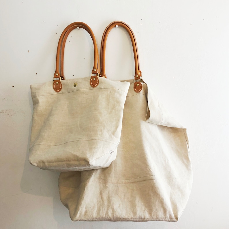 Puede incluir: Dos bolsas de lona beige con asas de cuero marr&oacute;n. La bolsa m&aacute;s peque&ntilde;a est&aacute; delante de la m&aacute;s grande.