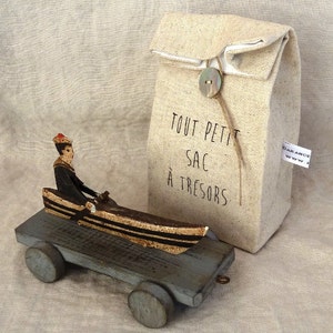 Op de afbeelding: Een houten speelgoedboot met een figuur van een persoon in een matrozenpak. De boot staat op een houten platform met wielen. De boot staat naast een beige stoffen tas met de tekst "Tout Petit Sac à Trésors" en een knoop.