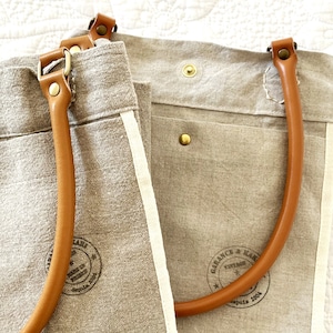 Puede incluir: Dos bolsas de lona beige con asas de cuero marr&oacute;n. La bolsa de la izquierda tiene un ribete blanco y un sello marr&oacute;n que dice "Garance &  Lark Vintage" y "Depuis 2004". La bolsa de la derecha tiene un asa de cuero marr&oacute;n y un bot&oacute;n de lat&oacute;n.