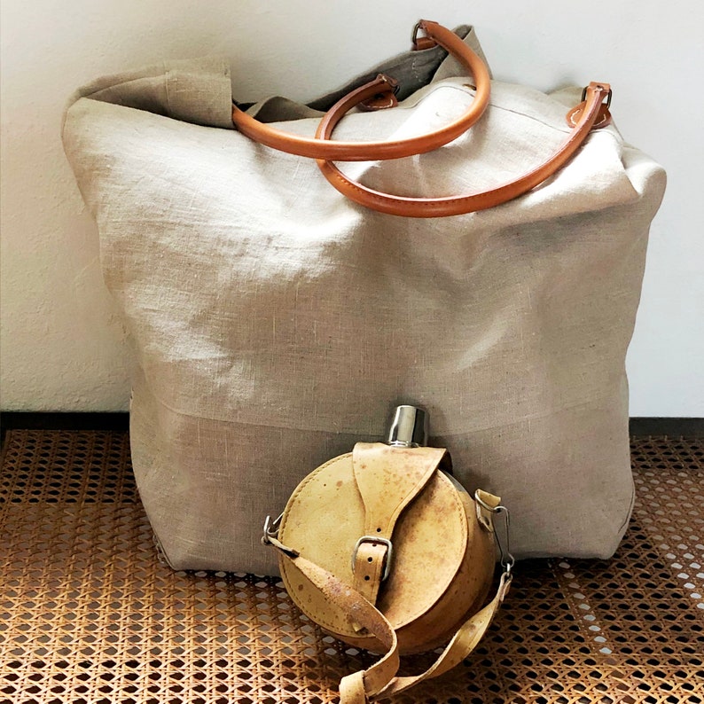 Puede incluir: Una gran bolsa de lona beige con correas de cuero marr&oacute;n y una cantimplora de cuero marr&oacute;n con tapa de metal.