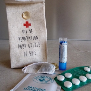Op de afbeelding: Een witte canvas tas met een rood kruis en de tekst "Kit de reparation pour gueule de bois". De tas bevat een klein wit zakje met een bruin theezakje, een blauwe tube en een groene blisterverpakking met witte pillen.