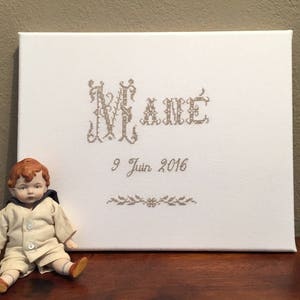 Peut inclure: Toile blanche avec le nom "Nolane" brodé en fil brun. La date "9 Juin 2016" est également brodée sous le nom. Une poupée en porcelaine aux cheveux roux et une tenue de marin blanche est assise devant la toile.